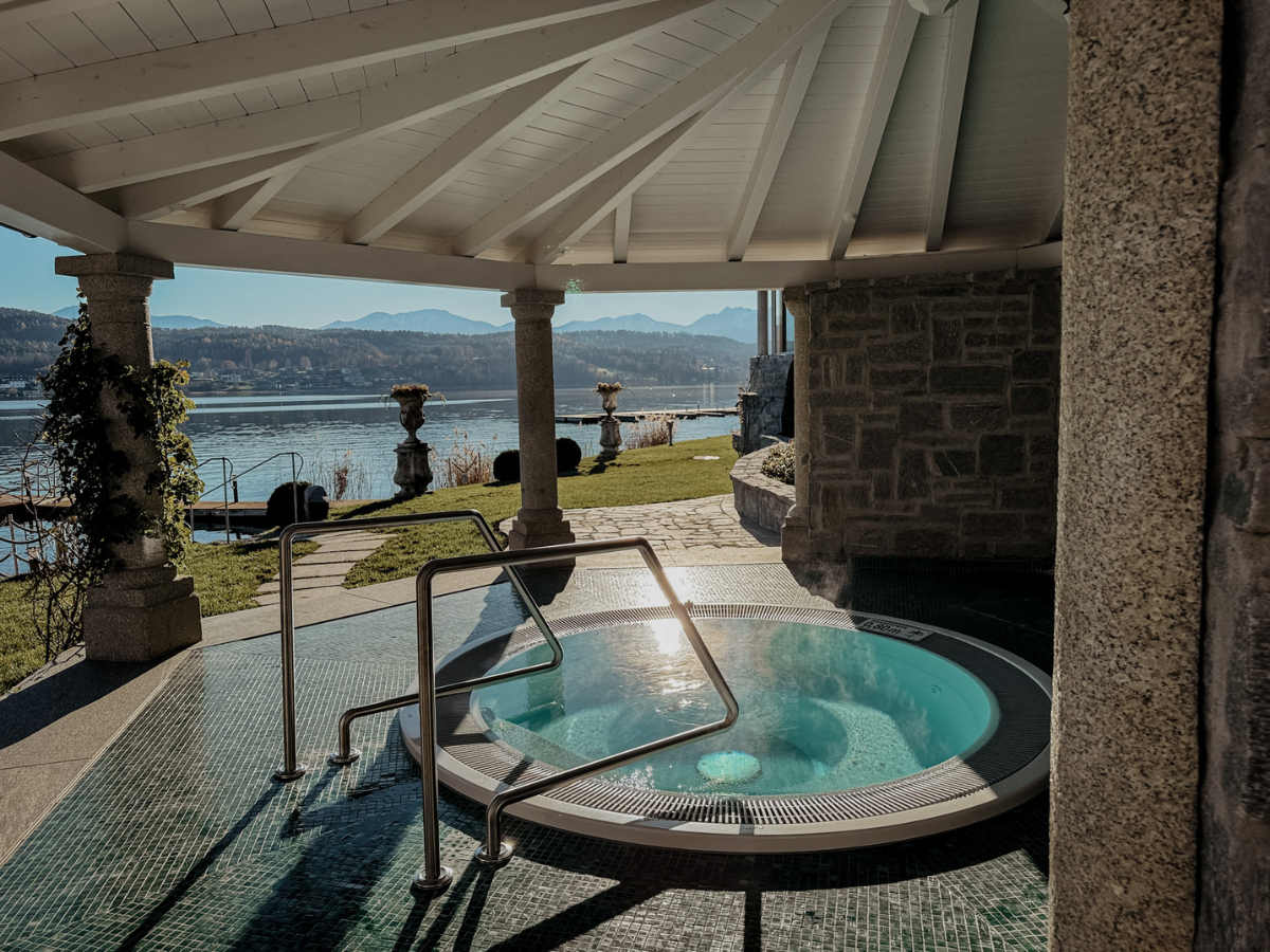 COCOtravel SchlossSeefels Spa Whirlpool1 Ein runder Whirlpool im Freien - der SchlossSeefels Spa Whirlpool - ruht unter einem weißen, offenen Pavillon mit Blick auf einen malerischen See und die Berge. Das Sonnenlicht glitzert auf dem Wasser, während Grünpflanzen und eine Steinmauer zu diesem ruhigen COCOtravel-Ausflug beitragen.